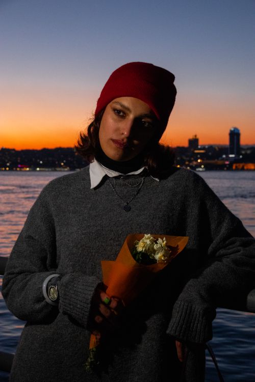 Free Woman in a red beanie holding white flowers at a waterfront during sunset. Stock Photo