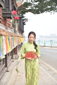A woman in a green traditional dress holding a red fan in a serene outdoor setting.