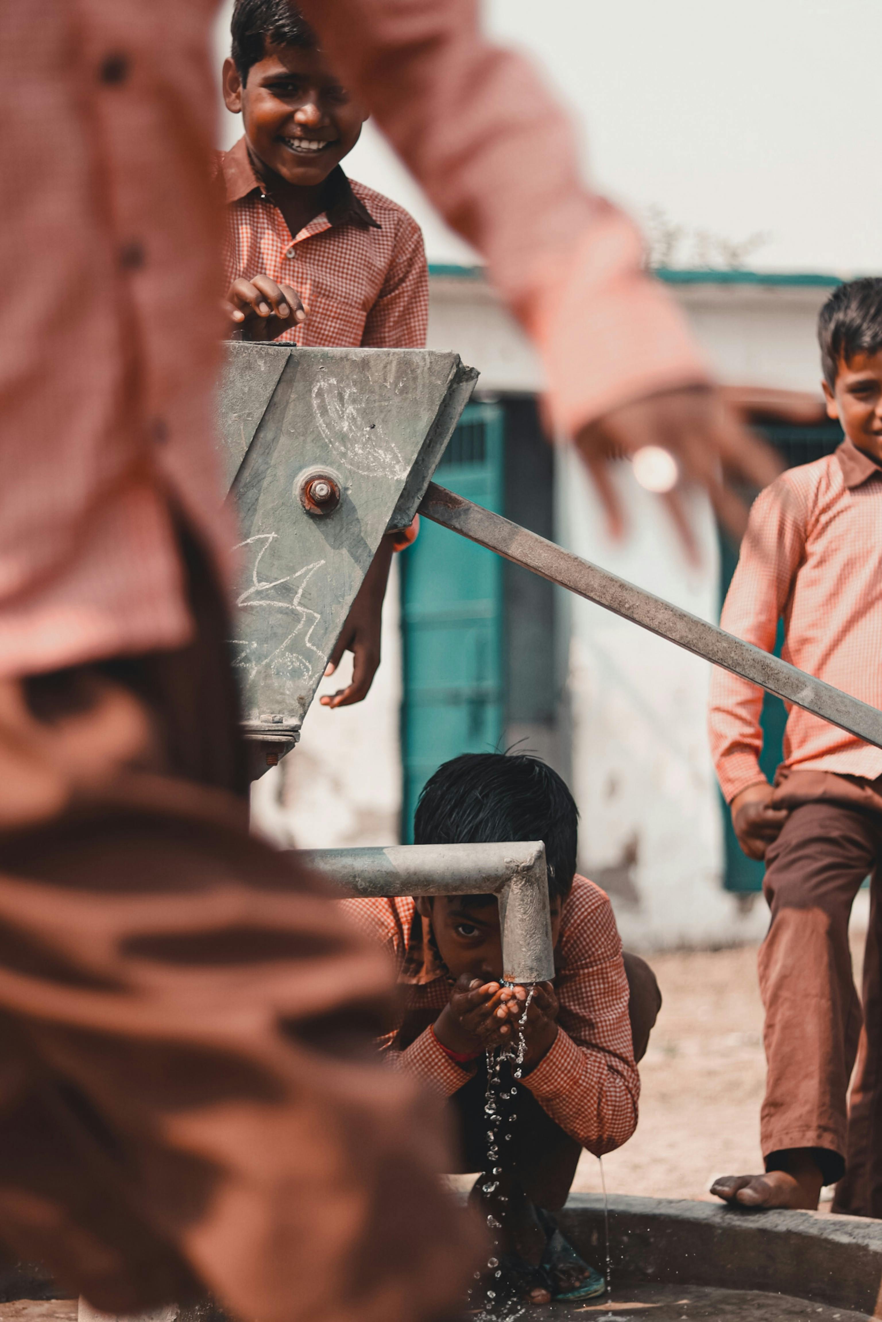 Group of boys drinking water from a hand pump outdoors, smiling and playful.