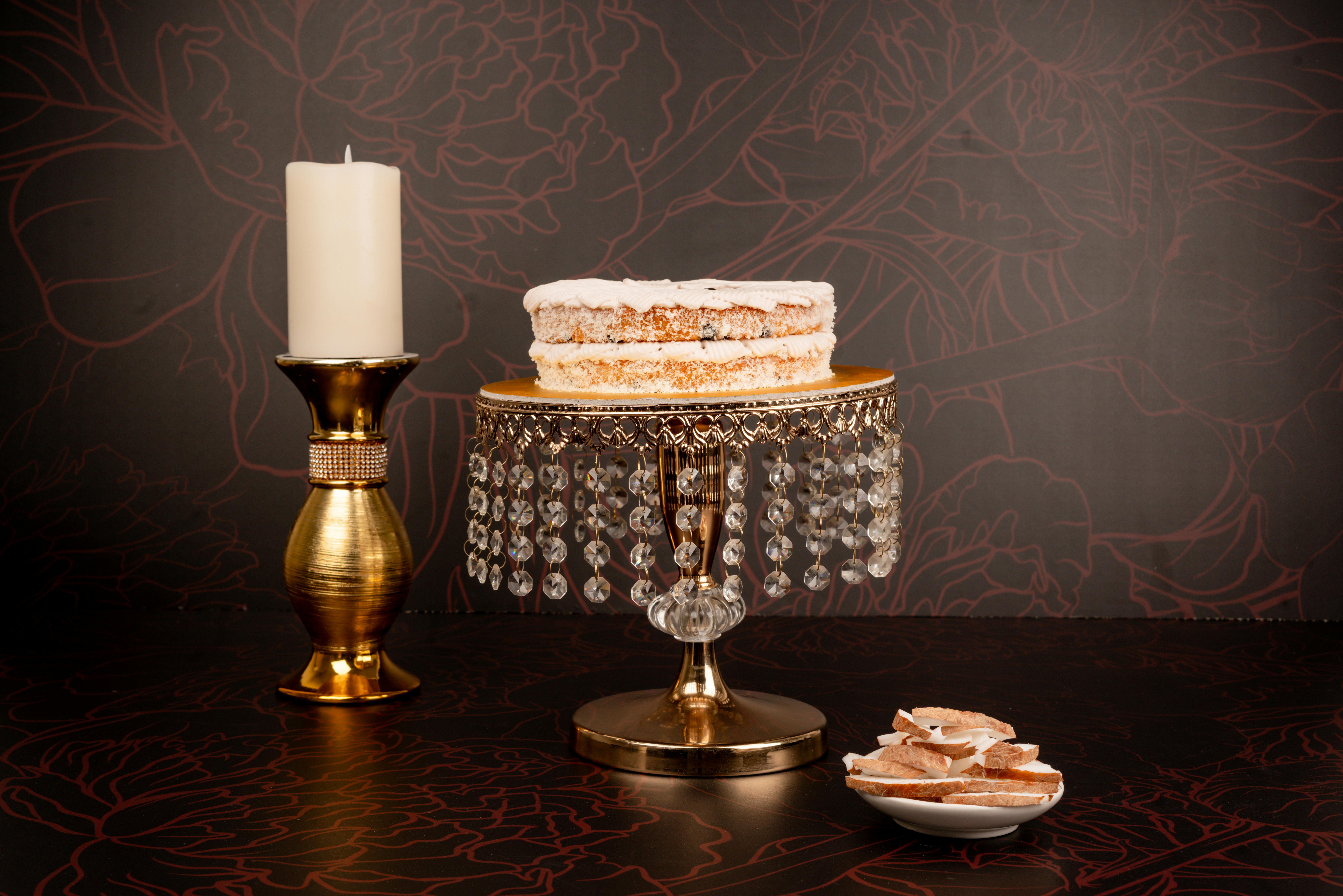 A beautifully iced cake on a crystal stand next to a candle, set against a floral backdrop.