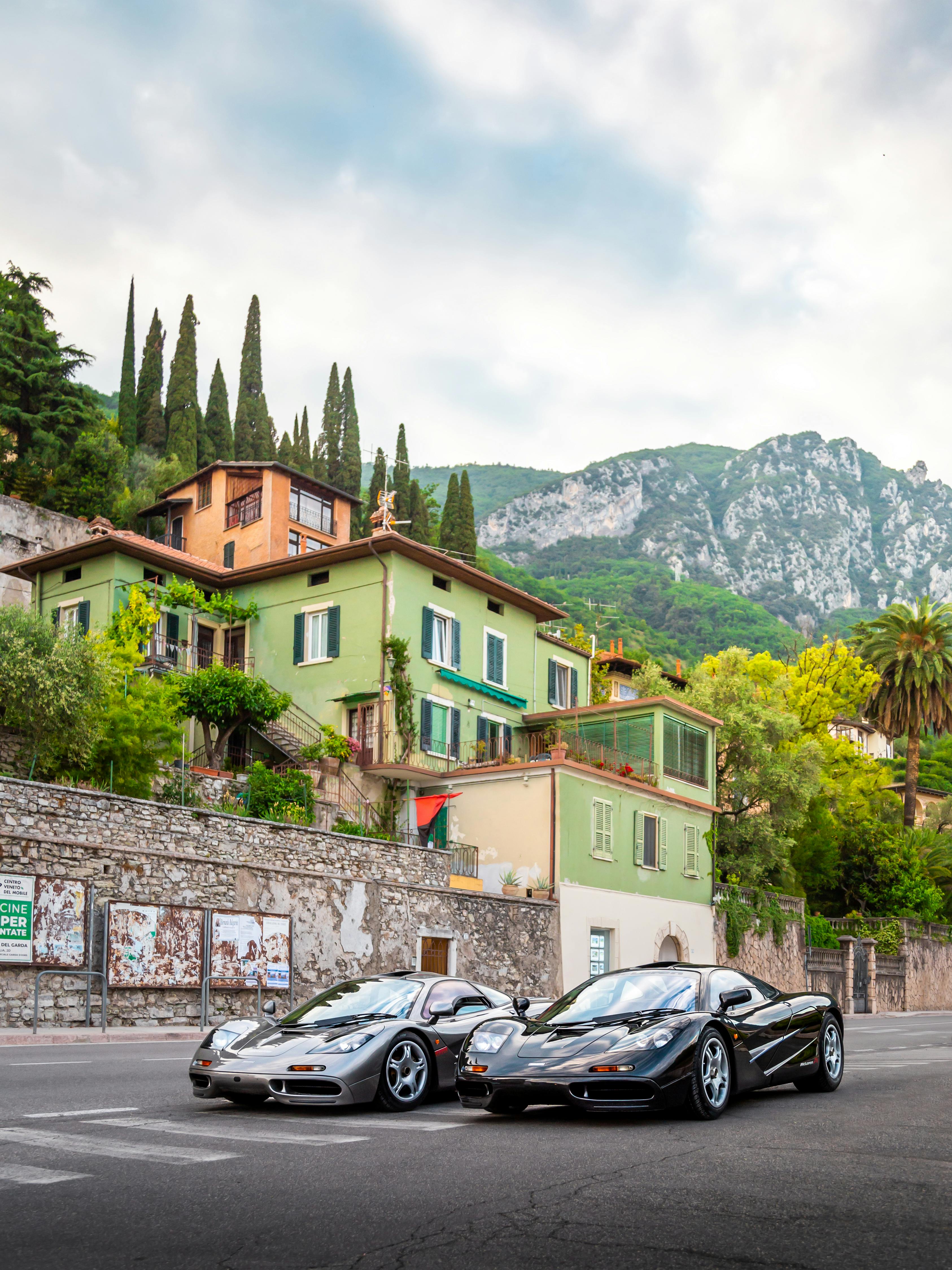 Two luxury sports cars parked in a scenic Italian Italian village with lush greenery and mountainous backdrop.