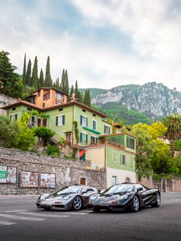 Two luxury sports cars parked in a scenic Italian village with lush greenery and mountainous backdrop.