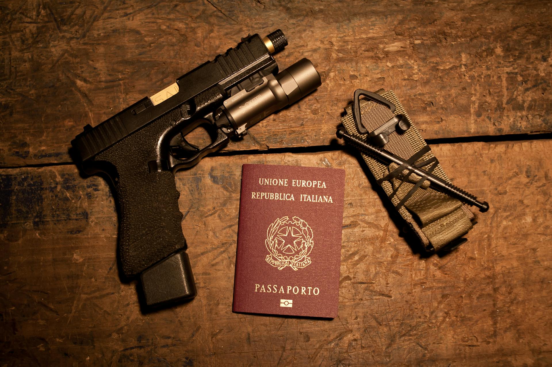 A gun, Italian passport, and tactical gear arranged on a wooden surface, hinting at travel or security themes.