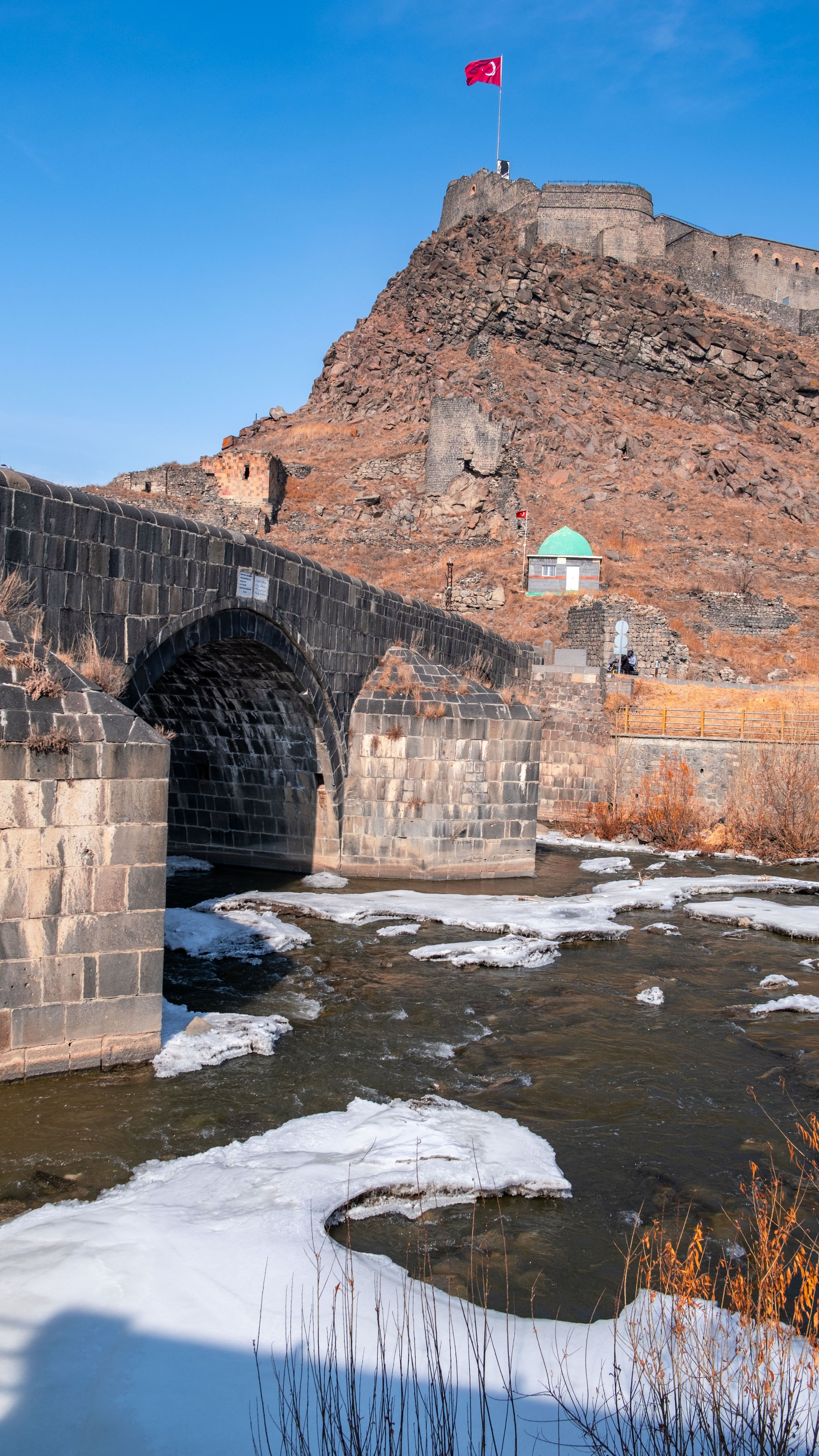 View of Kars Castle and a stone bridge over a partially frozen river in Türkiye.