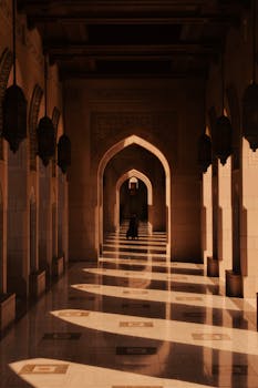 Warm sunlight casts intricate shadows in a corridor of the Sultan Qaboos Mosque, Muscat, Oman.
