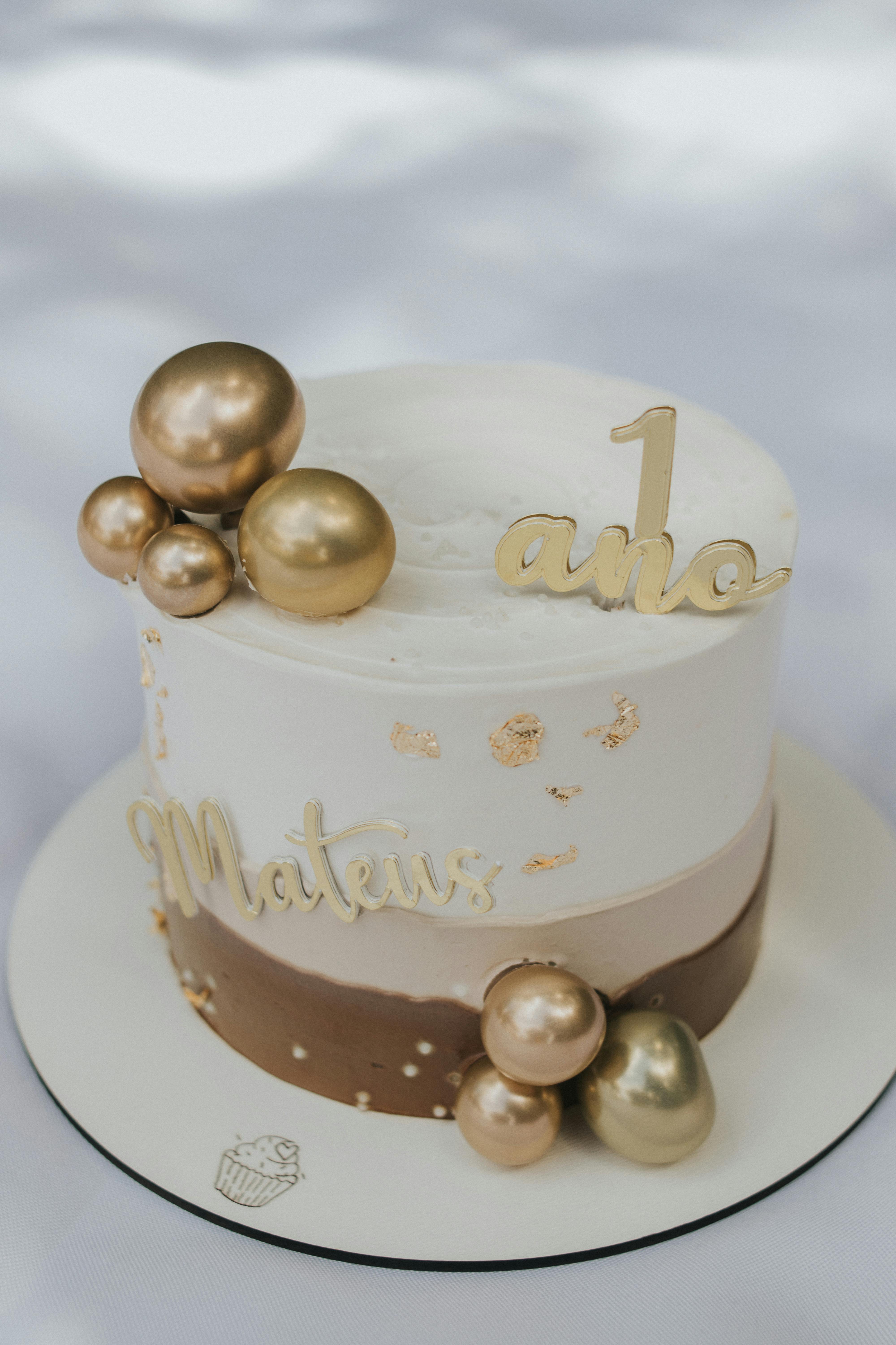 Elegant gold and white birthday cake with decorative spheres and '1 ano' topper.