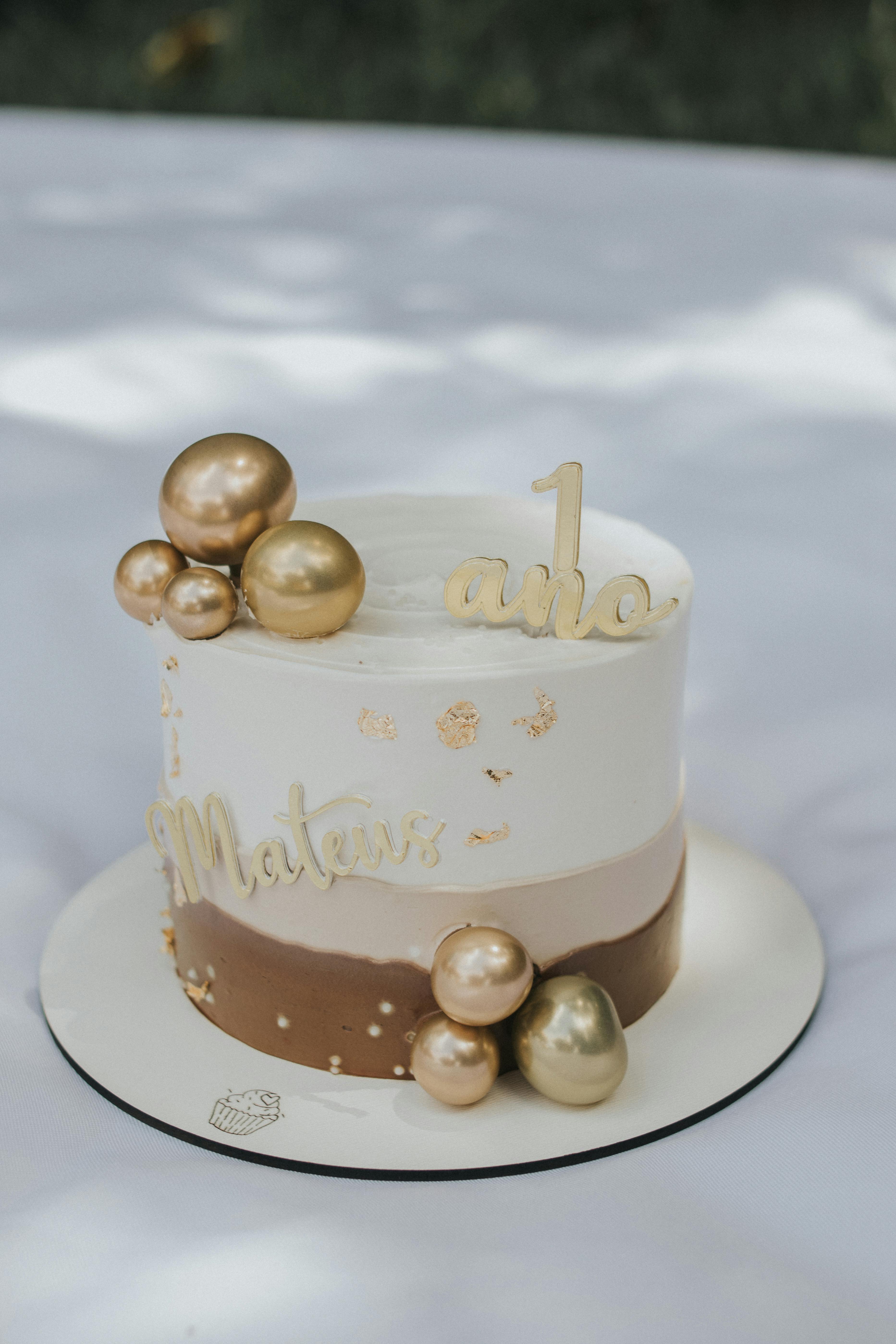 White and gold first birthday cake with decorative spheres and topper.