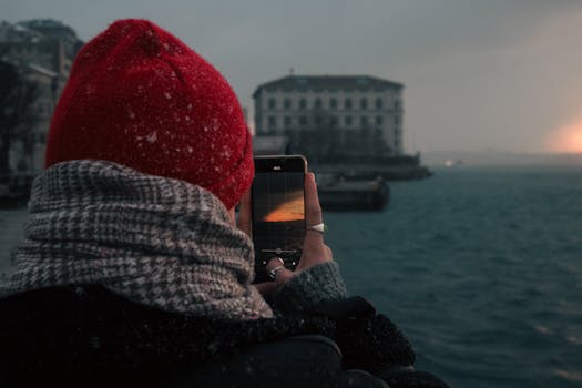 A person captures a scenic waterfront view in winter twilight with their smartphone, wrapped warmly.