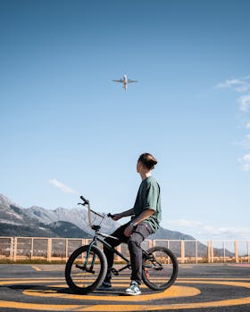 A young man on a BMX bike observes a plane flying overhead on a sunny day with clear blue skies.