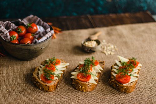Free Rustic multigrain bread topped with cheese, tomatoes, and dill on a burlap surface, perfect for food photography. Stock Photo