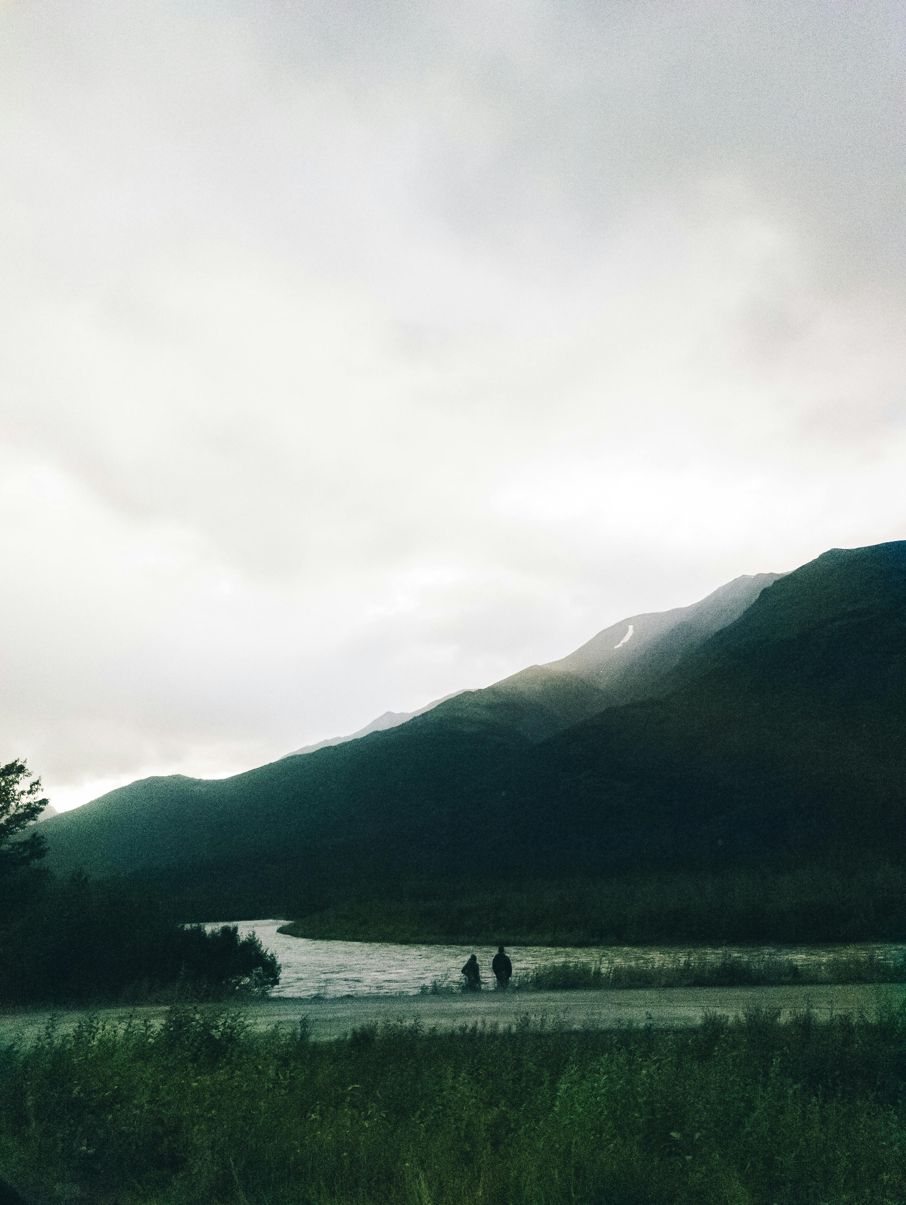 Two people fishing by a river in the scenic Alaskan wilderness with mountainous backdrop.