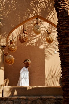 A man in traditional attire stands near an arched doorway in Nizwa, Oman, casting shadows.