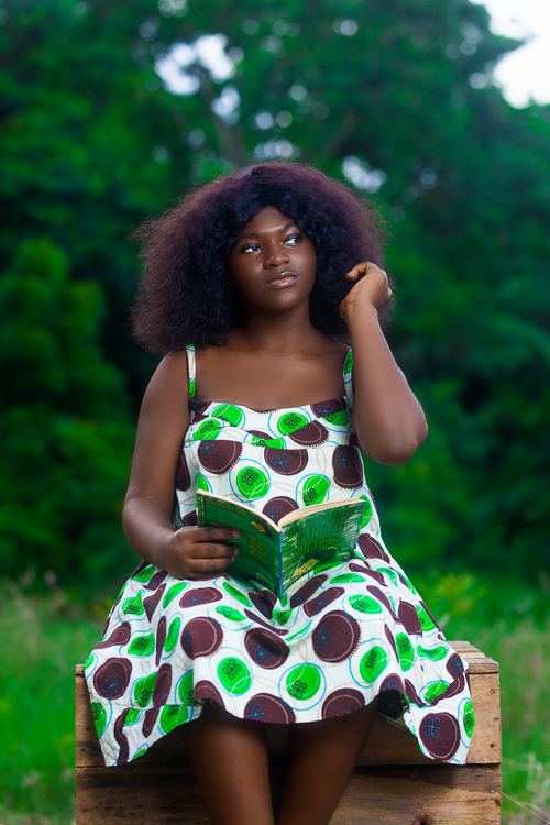 Free Young woman sits outdoors reading a book against a lush green background. Stock Photo