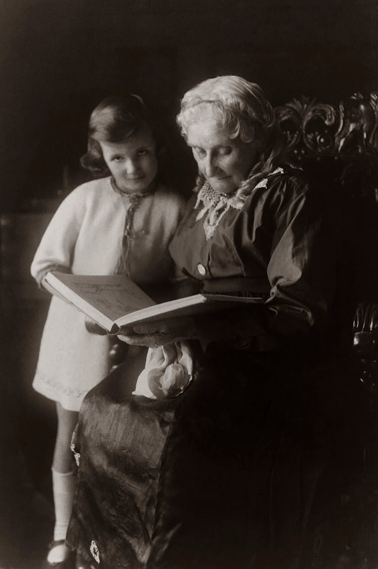 Old Woman Sitting On Chair And Reading A Book To A Child