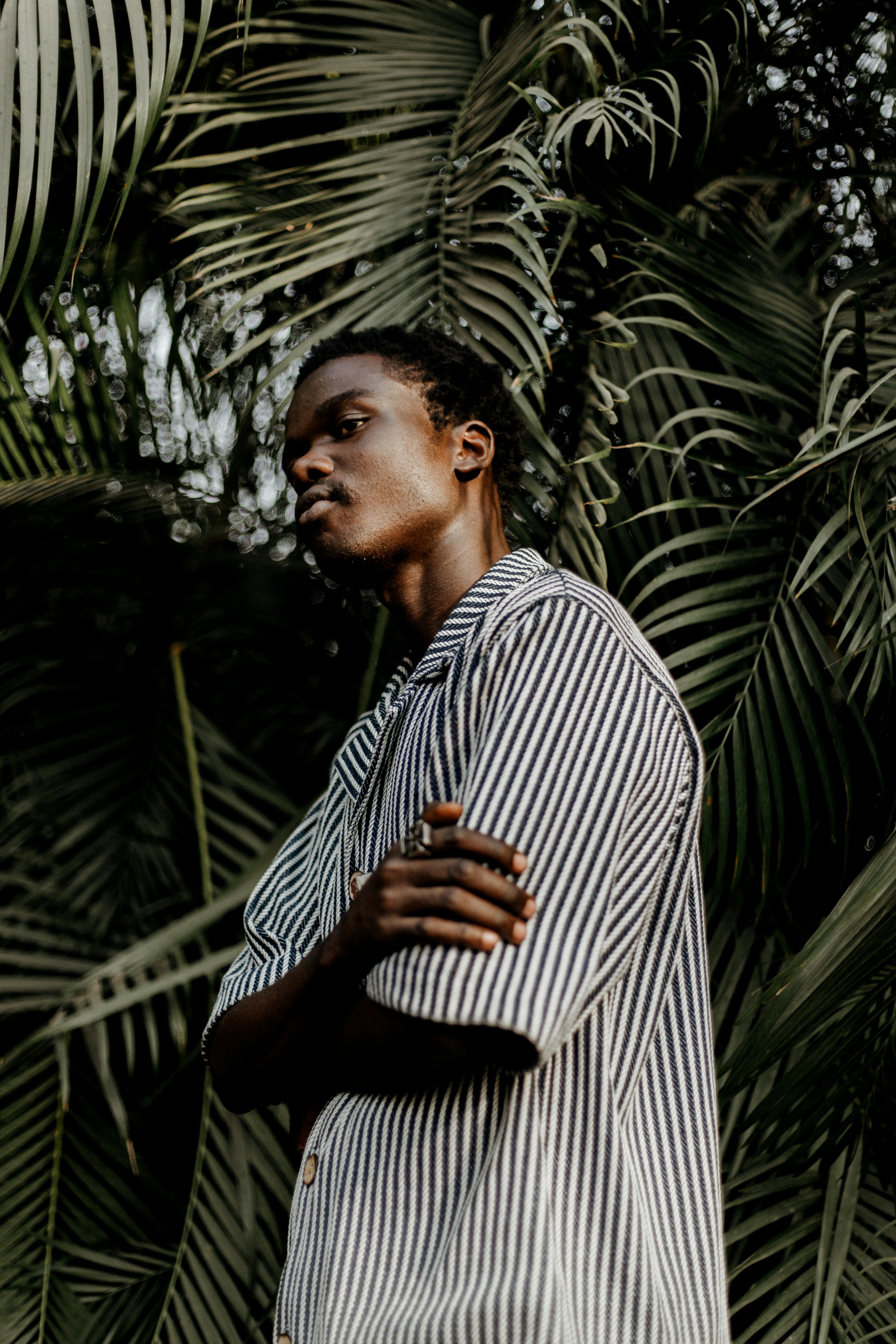 Stylish man in striped shirt stands confidently before lush plant background.