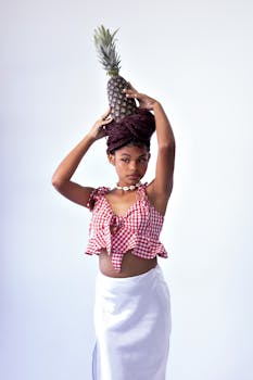 Young woman in red checked top balancing a pineapple gracefully on her head in a studio setting.