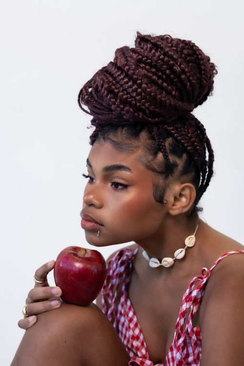 Free A young woman with braided hair holds a vibrant red apple, exuding a playful yet thoughtful aura. Stock Photo