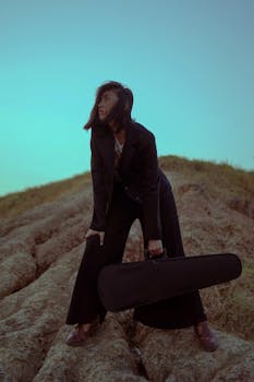 一位时尚女子在日落时分，身着黑色西装，手持吉他盒，置身于自然场景中