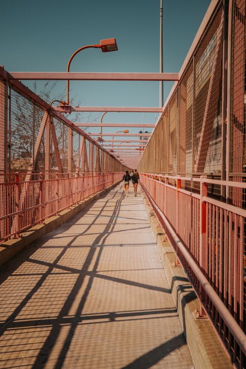 Free Two people walking on a sunny bridge pathway in New York City, showcasing urban architecture. Stock Photo