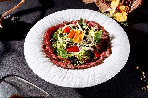 Free High-angle shot of gourmet beef carpaccio garnished with fresh greens and edible flowers on a textured plate. Stock Photo