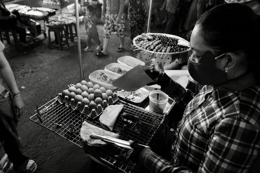 Masked street vendor grills eggs at a bustling night market, creating a lively scene.