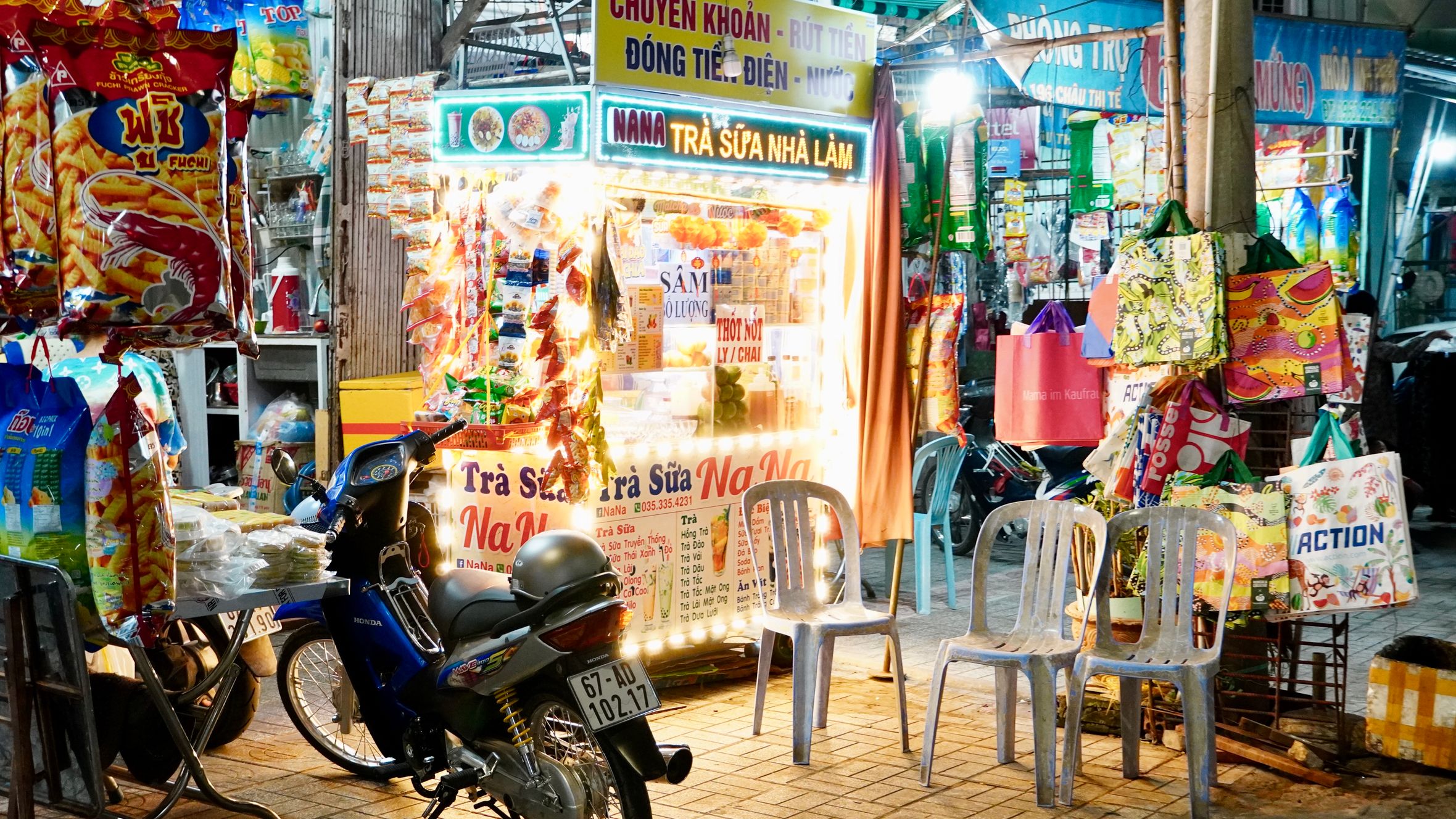 Colorful street vendor stall at night market with hanging snacks and plastic chairs, Vietnam.