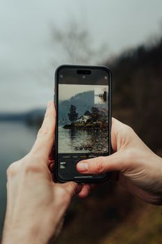 A person taking a scenic lake photo with a smartphone on a cloudy day.