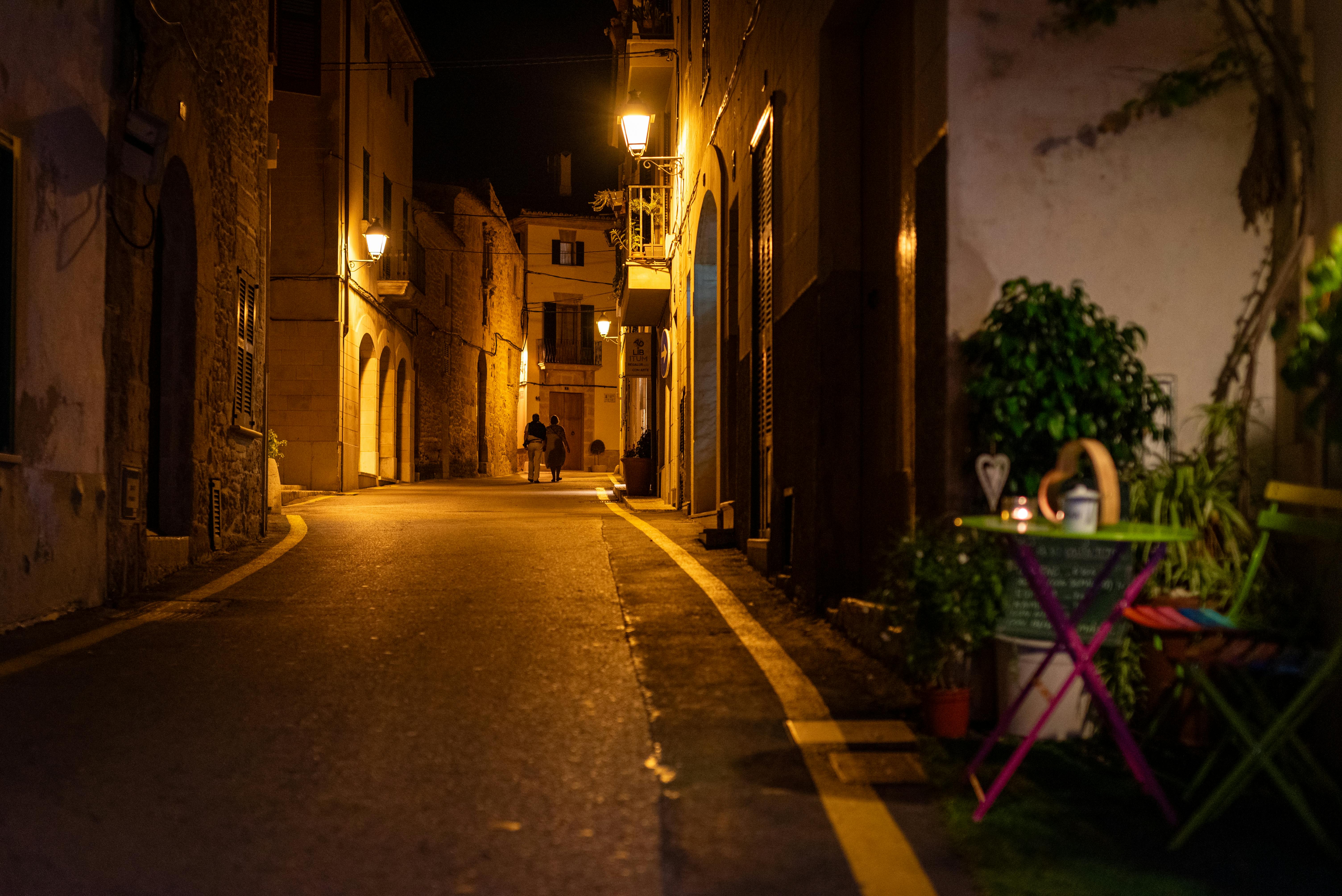 Couple enjoying a romantic evening walk on a charming narrow street in Alcúdia, Spain, lit by warm streetlights. - Alcúdia