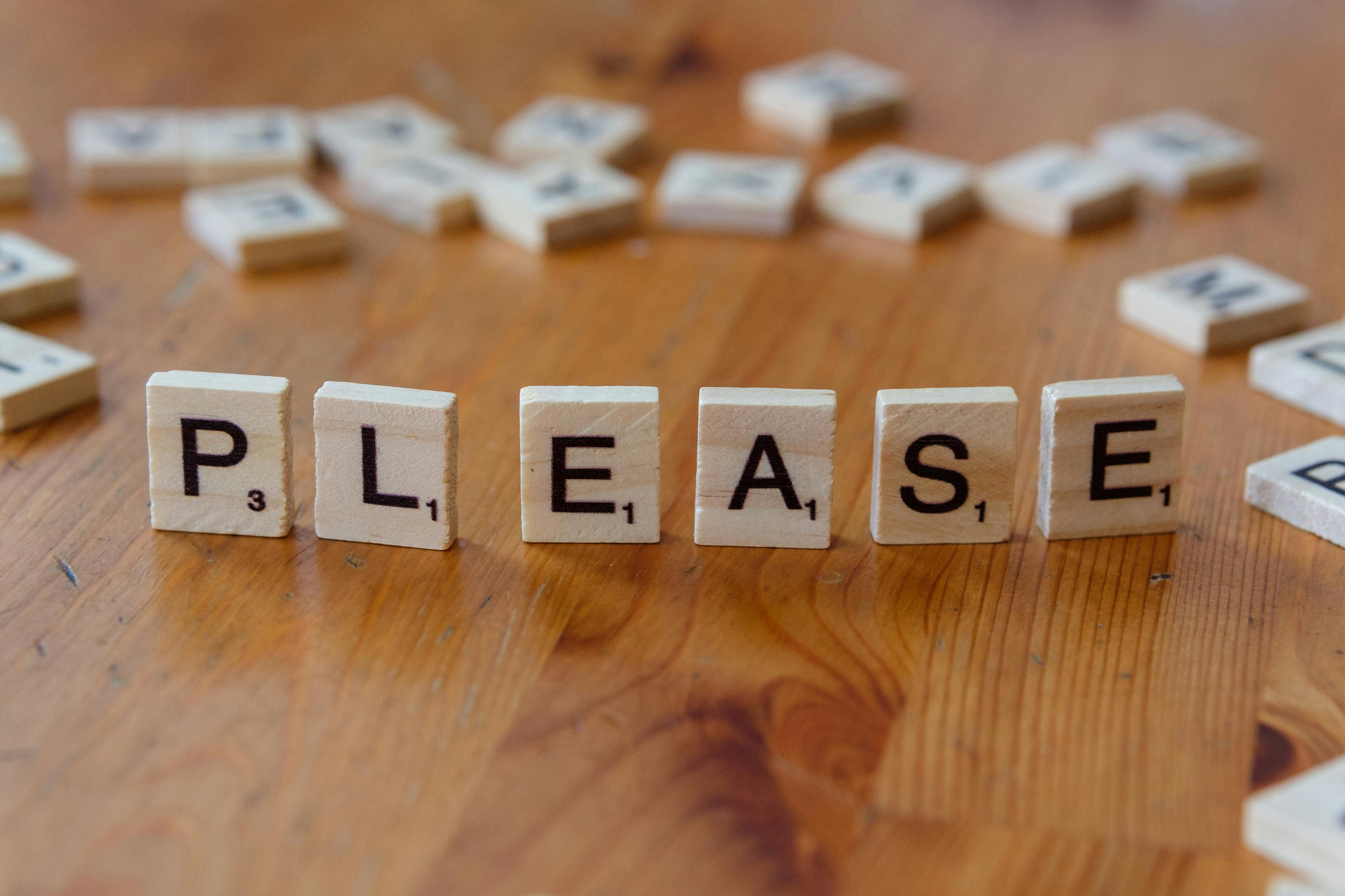 Free Scrabble tiles arranged to spell 'Please' on a wooden surface, symbolizing politeness. Stock Photo