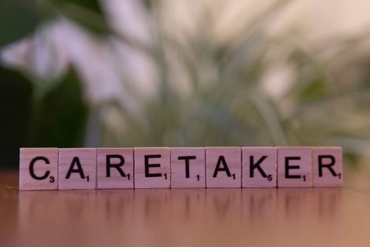 Close-up of Scrabble tiles spelling 'caretaker' on a wooden surface with blurred green background.