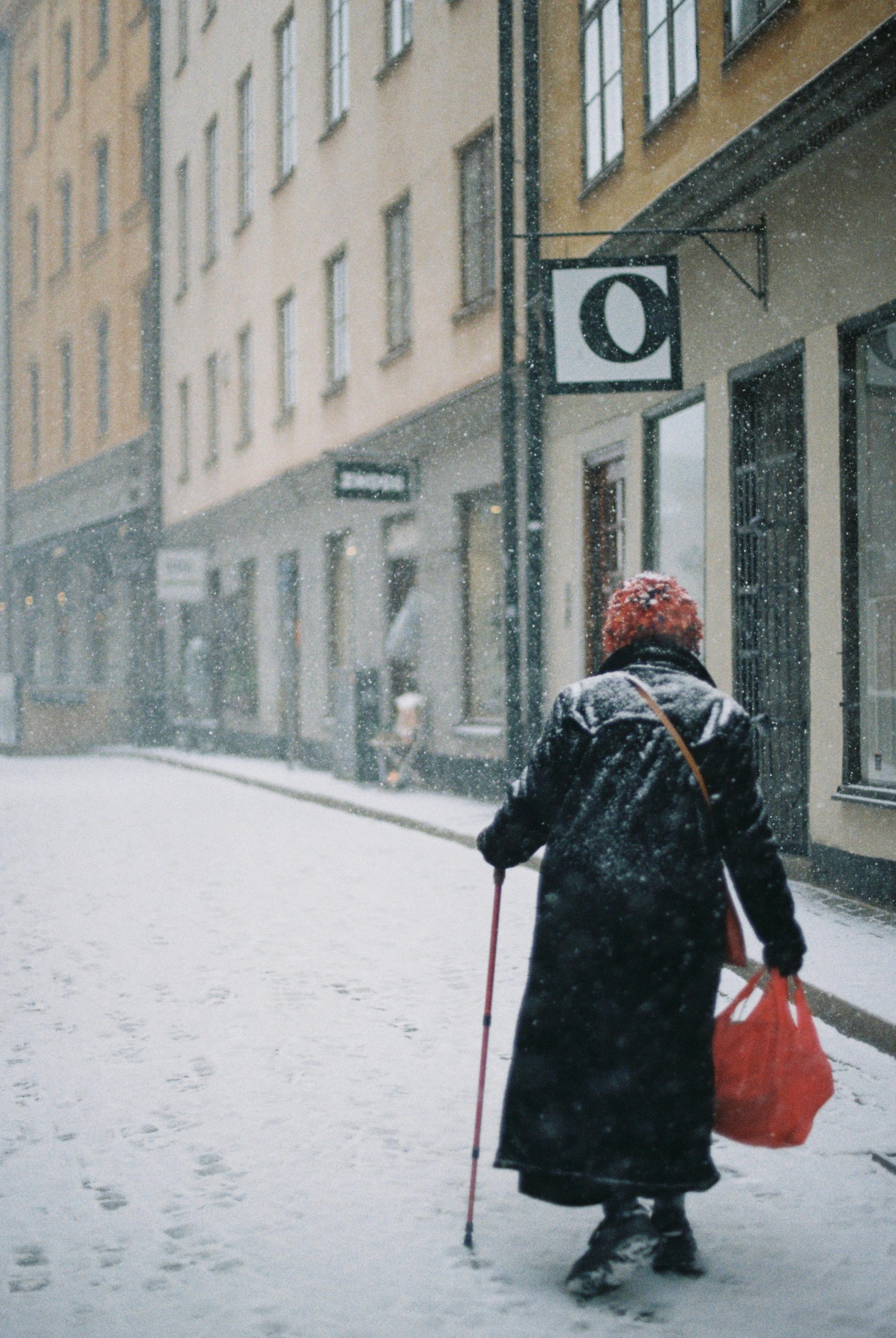 Elderly person walks through a snowy city street holding a cane and red bag during winter.