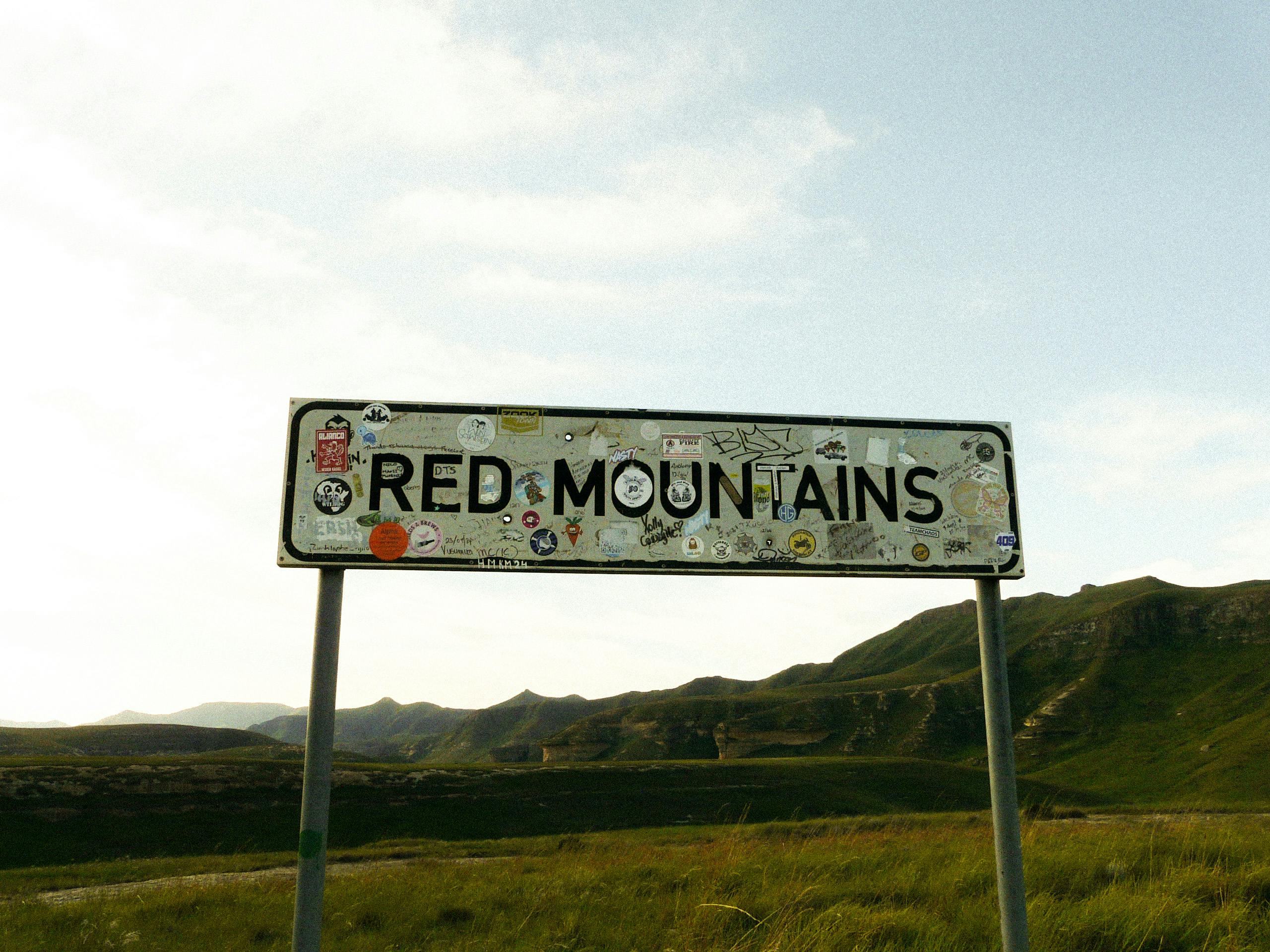 Outdoor view of a sign marking the Red Mountains with a backdrop of green hills and clear sky.
