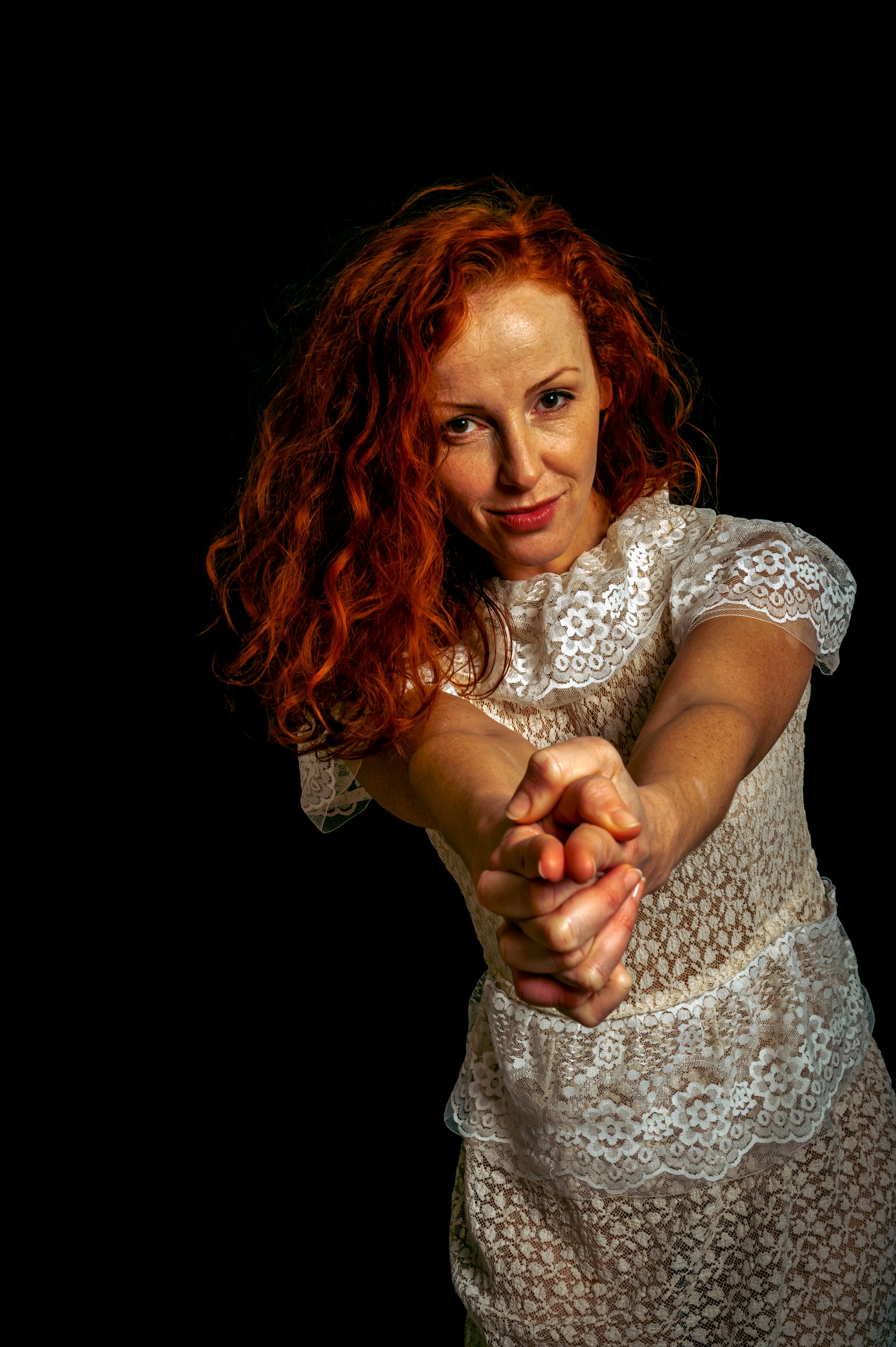 Portrait of a red-haired woman confidently posing with hands extended against a dark background.