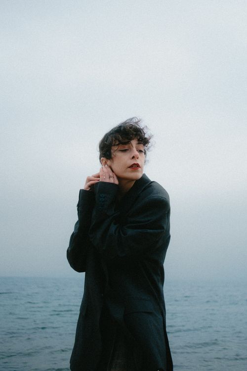 Free A contemplative woman in a coat stands by a calm sea under an overcast sky. Stock Photo