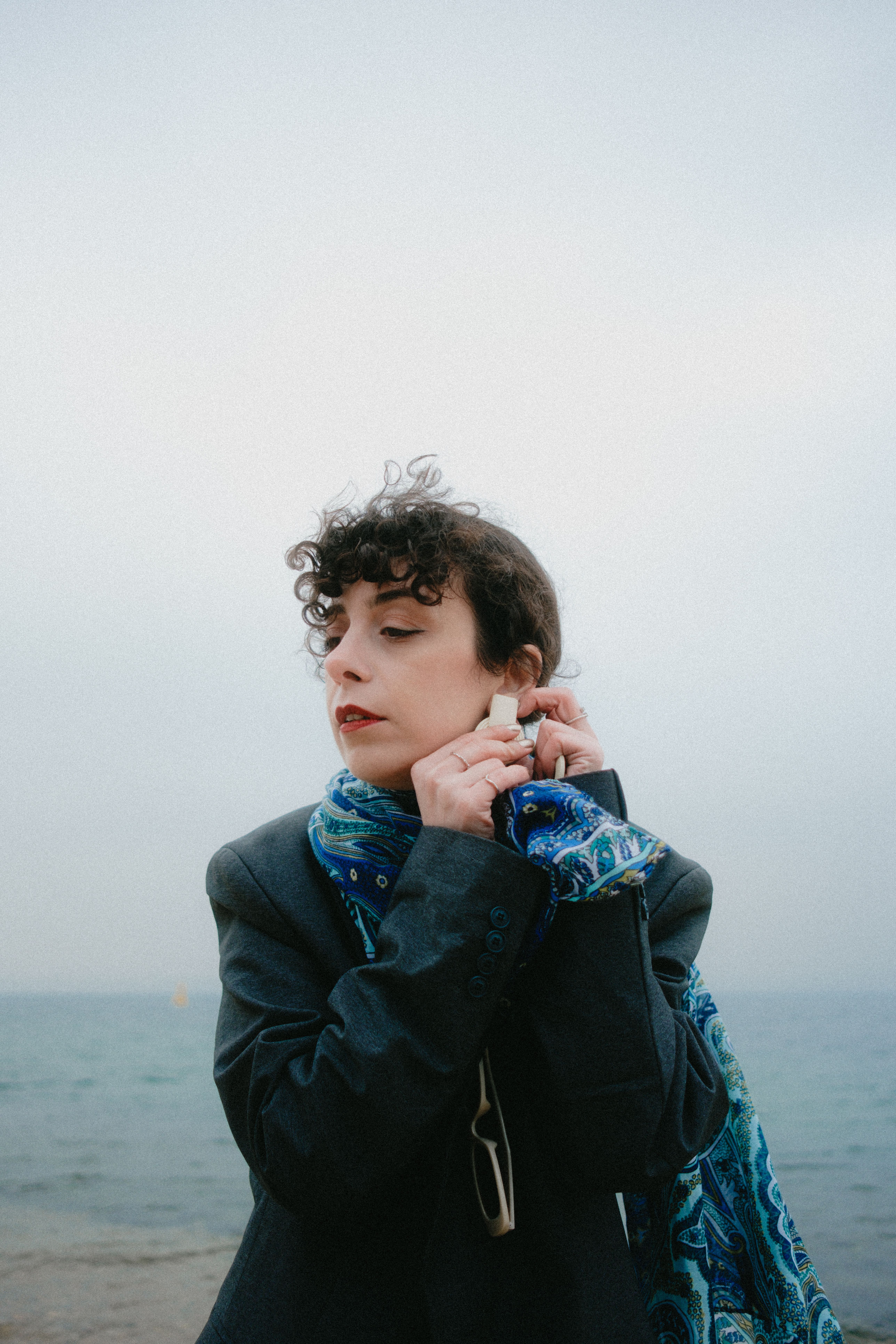 Stylish woman adjusts earring near the ocean, embracing vintage fashion with a blue scarf.