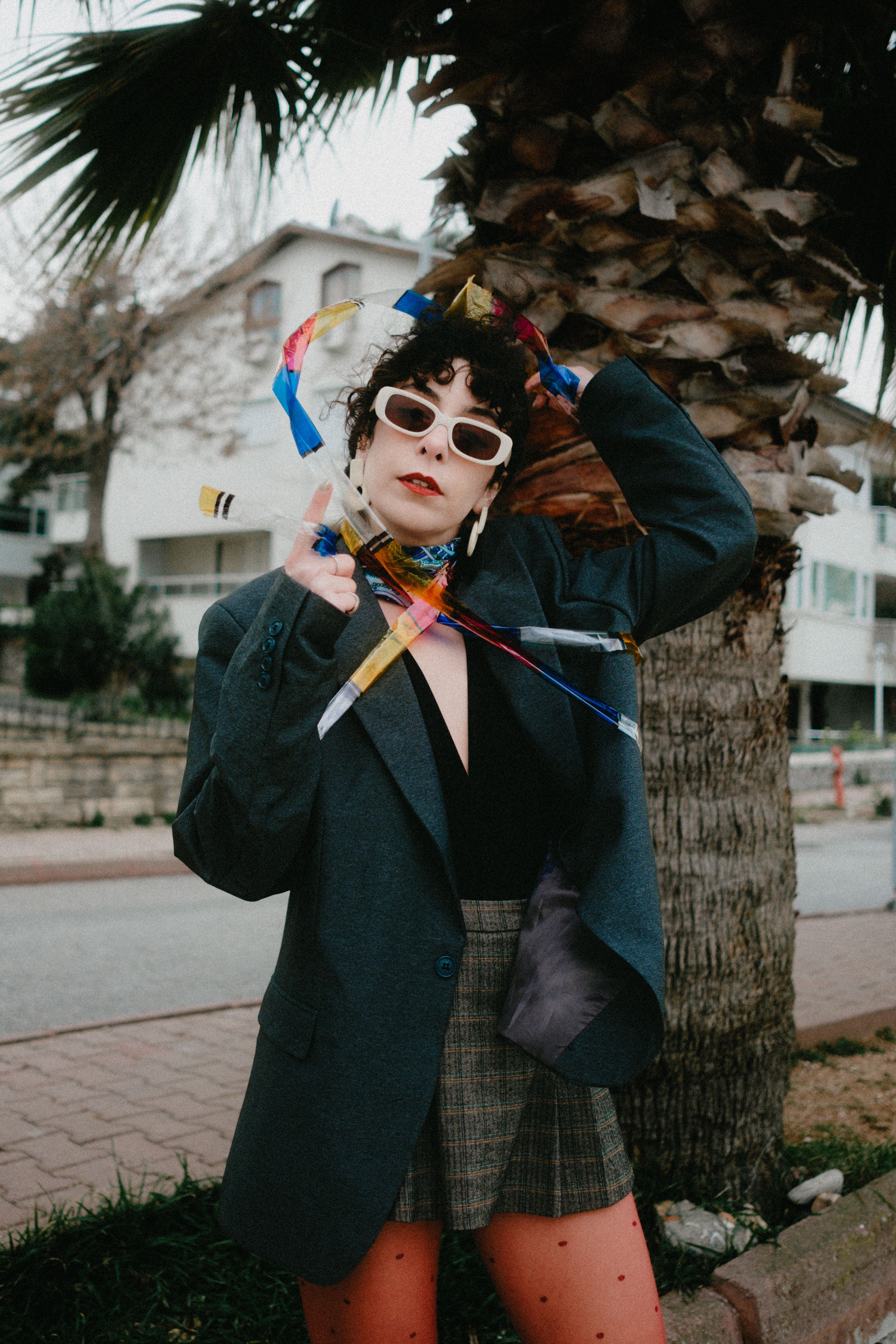 Stylish woman poses with colorful ribbons and oversized blazer outdoors by a palm tree.