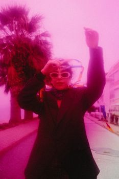 Artistic portrait of a person wearing bright accessories with a pink hue background and palm trees.