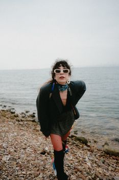 Stylish woman in retro outfit and sunglasses poses by the rocky seaside.