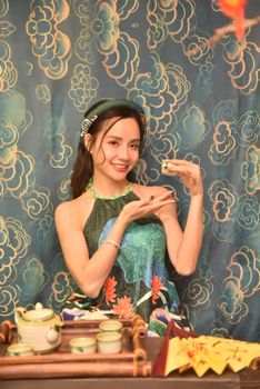Asian woman in a traditional dress enjoying a tea ceremony with intricate background.