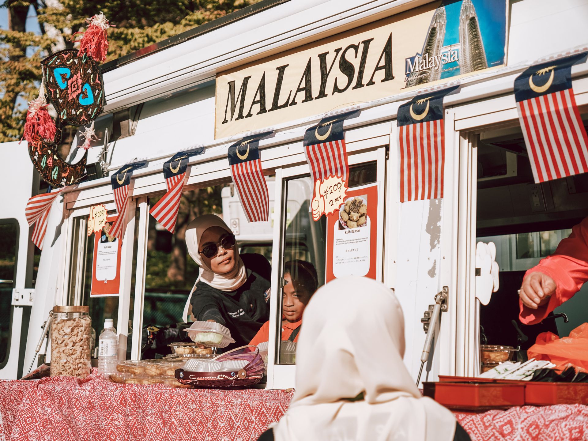 A vibrant Malaysian street food vendor serving customers at a market stall with traditional flags.