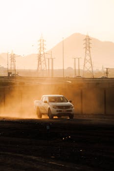 A white truck navigates a dusty road in Umman with mountains and power lines in the background.