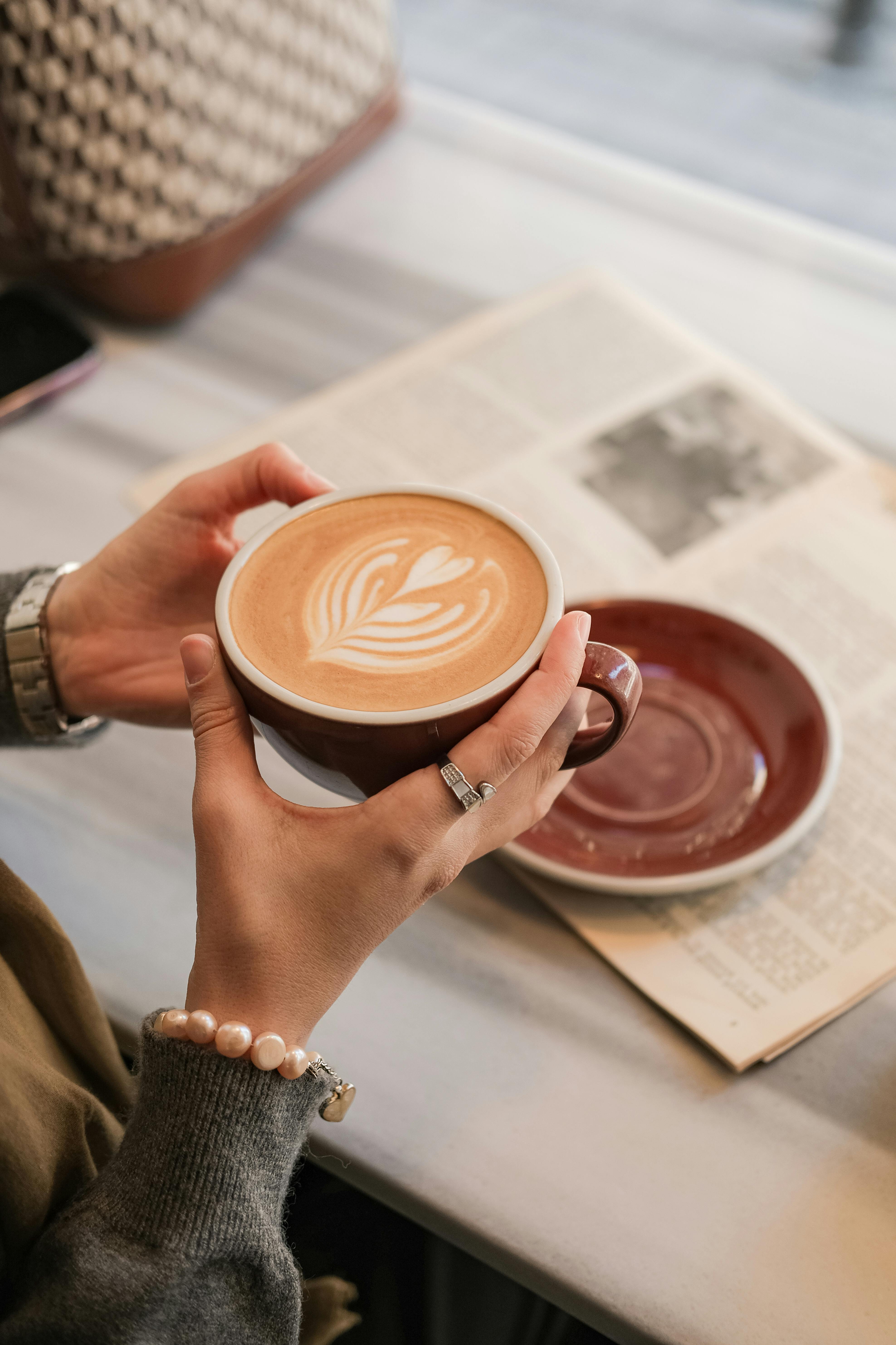 Hands holding a latte with artistic foam design, next to a newspaper on a cafe table.