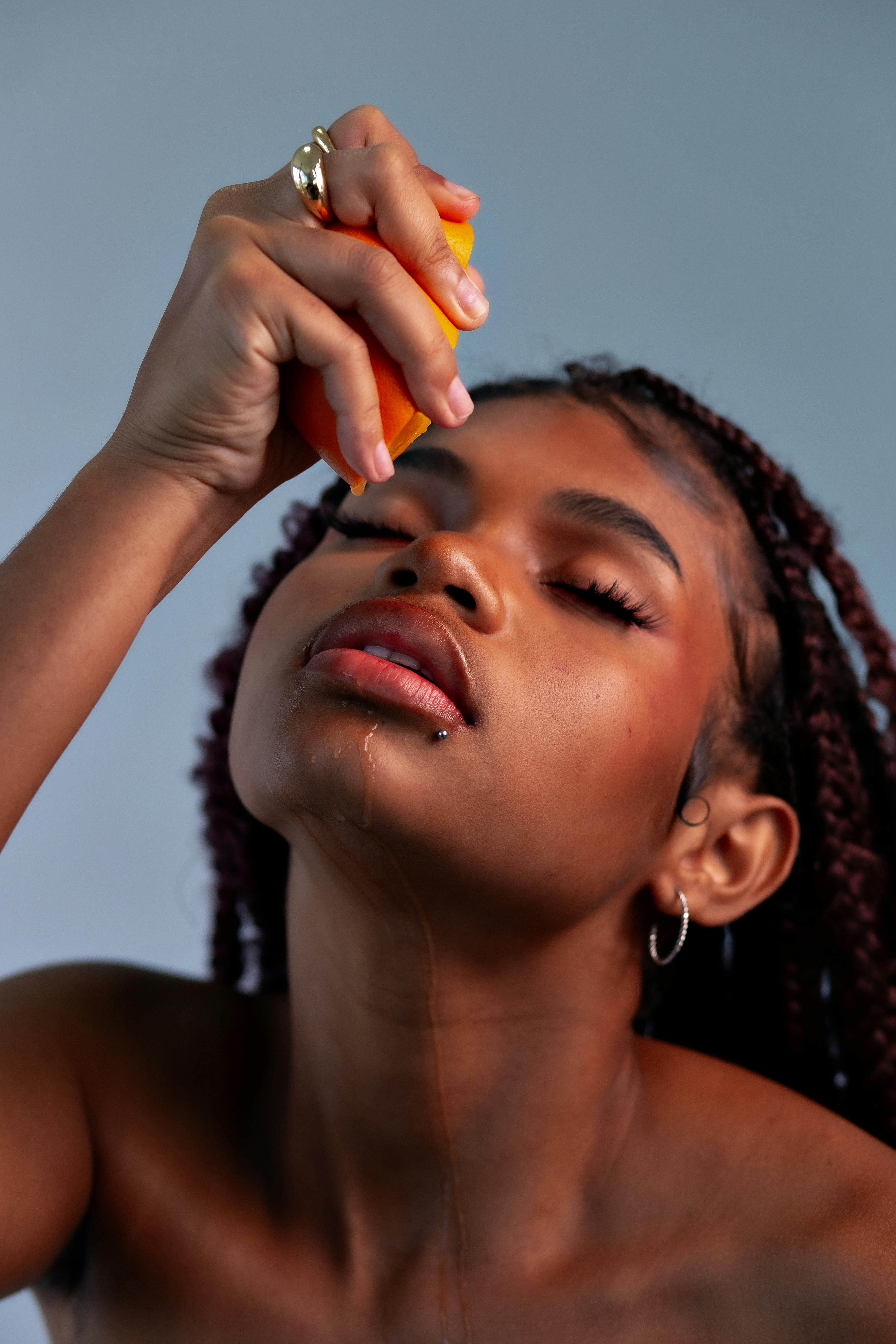 A young woman squeezing fresh orange juice onto her face with relaxed expression.