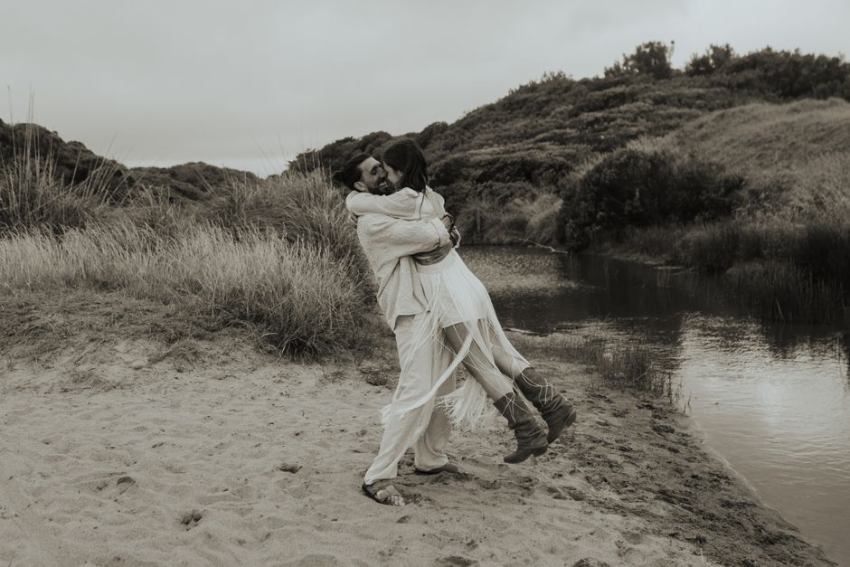 A couple embracing joyfully on a sandy beach with scenic hills in the background.