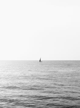 A solitary sailboat on a vast sea under a serene sky, captured in black and white.