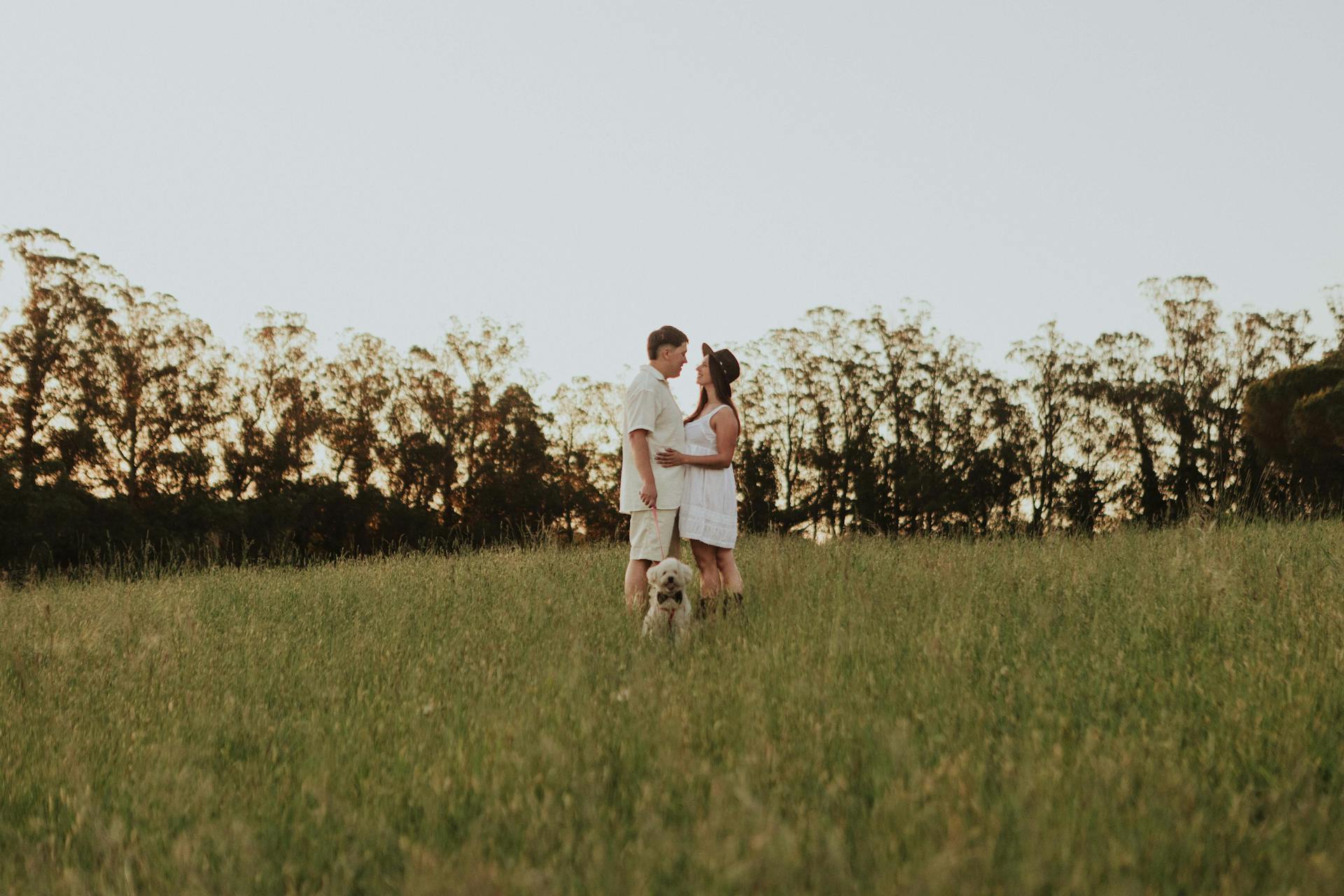 A couple embracing at sunset with their dog in a serene grassy field.