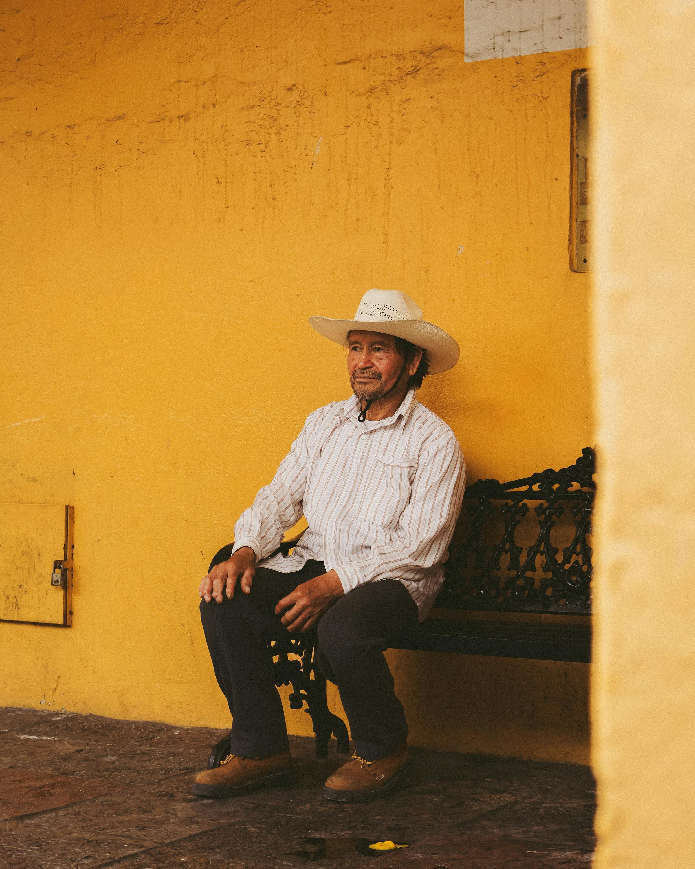 Senior man in traditional hat sitting on a bench against a vibrant yellow wall in Puebla, Mexico.