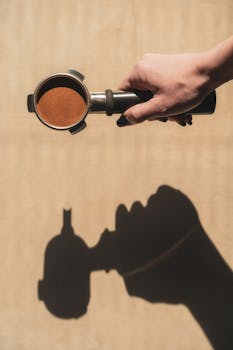 A hand holds a coffee portafilter casting a shadow on a light surface, creating a modern aesthetic.