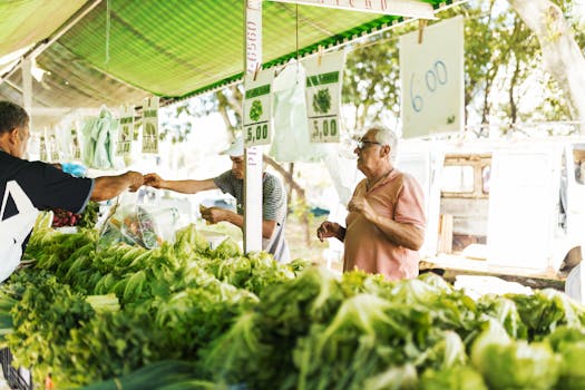 Customers purchasing fresh vegetables at an outdoor farmers market under green canopy.