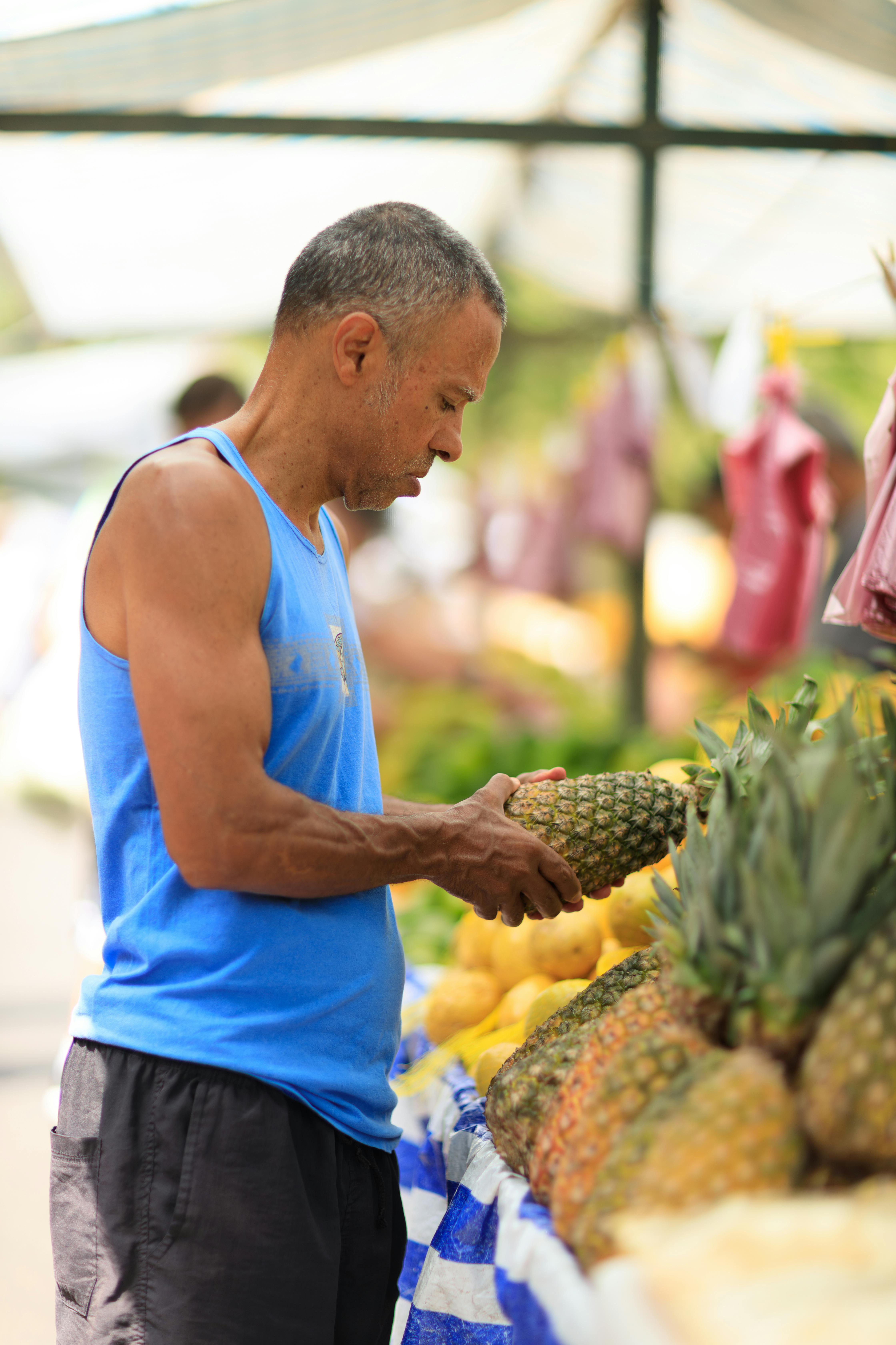 Adult man in blue tank top picks pineapples at an outdoor market, exemplifying healthy lifestyle choices.